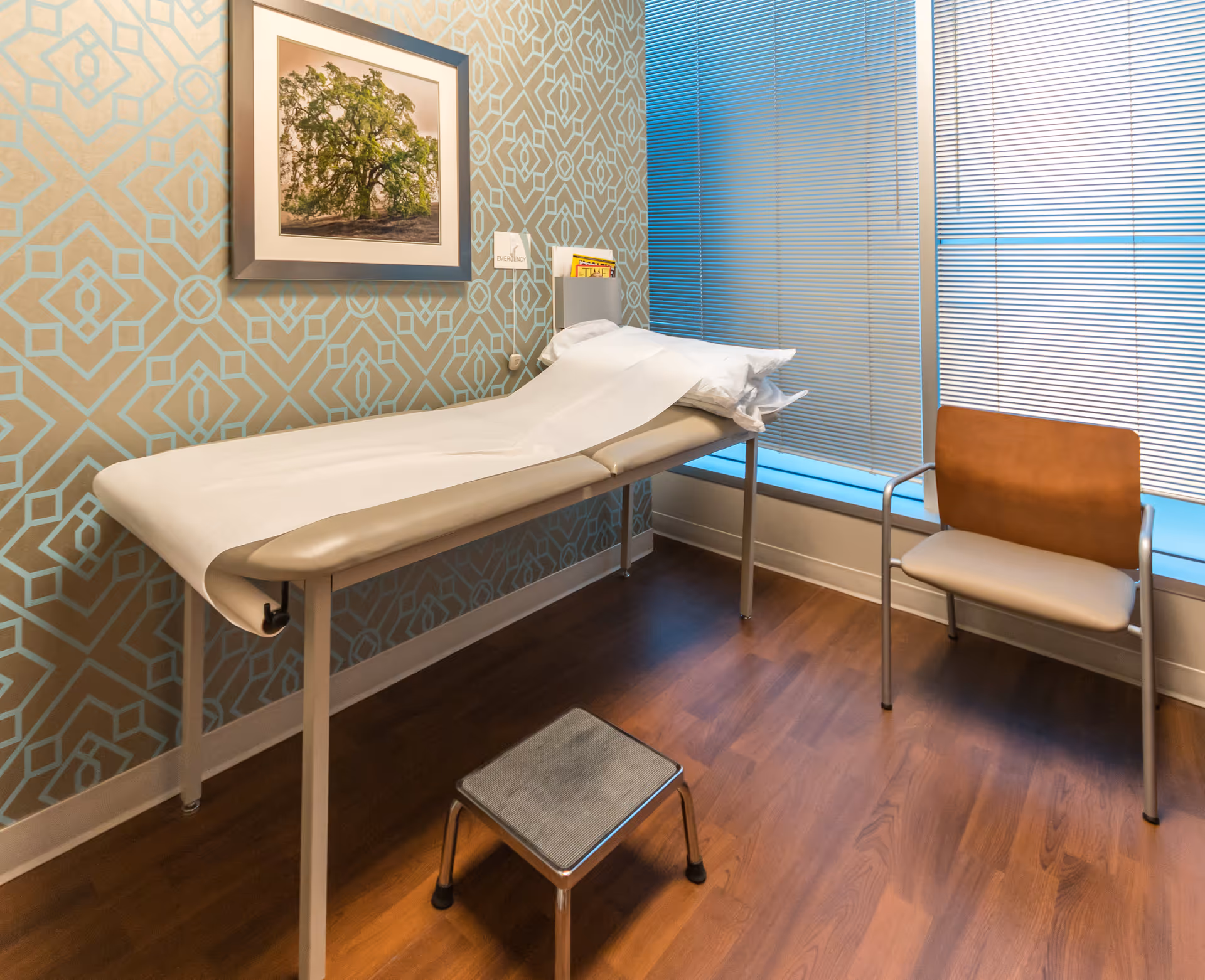 Medical examination bed in a small room with blue geometric wallpaper and blinds.