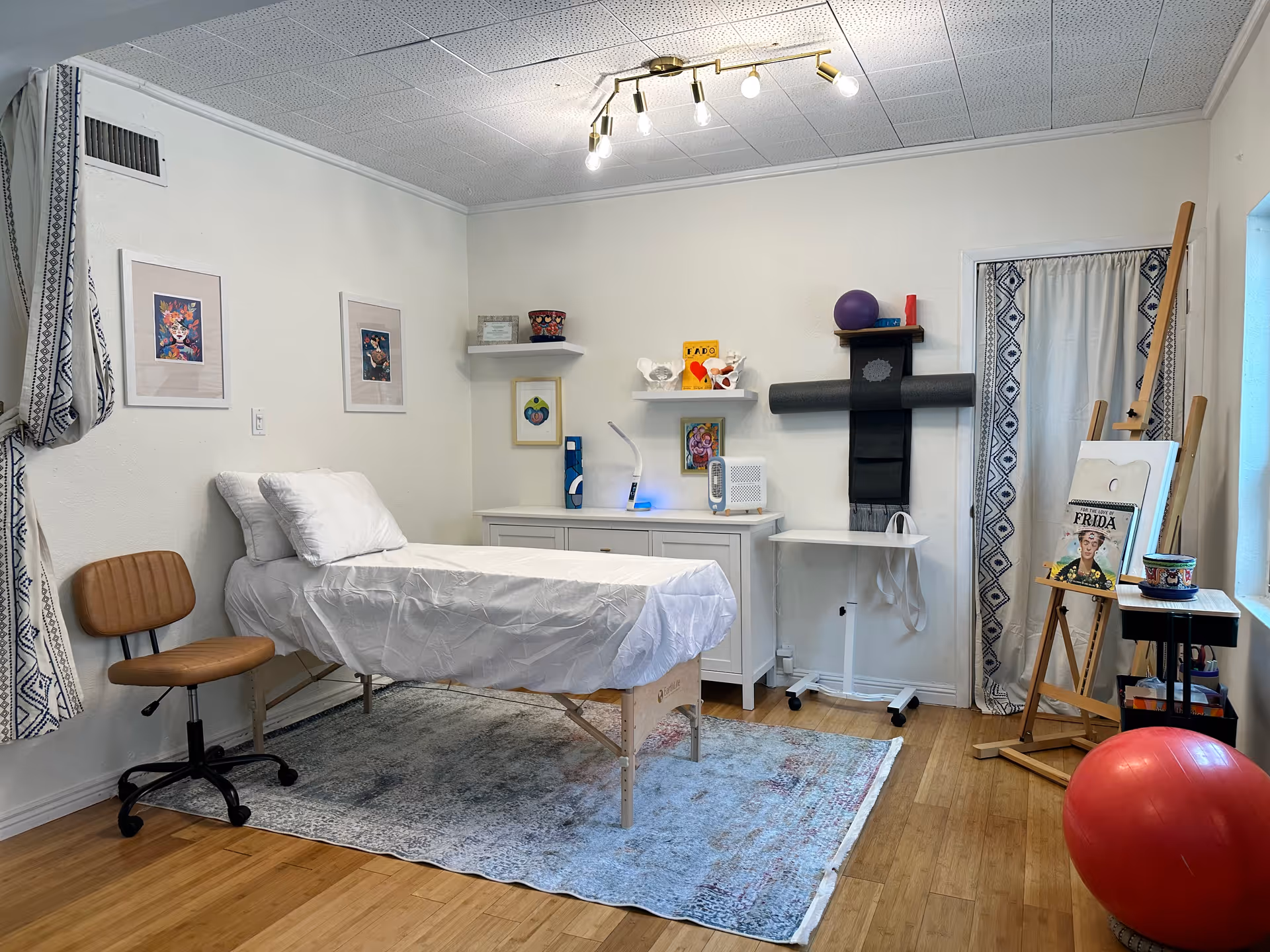 Therapy room with a bed, chair, cabinets, shelves, exercise ball, and curtains.