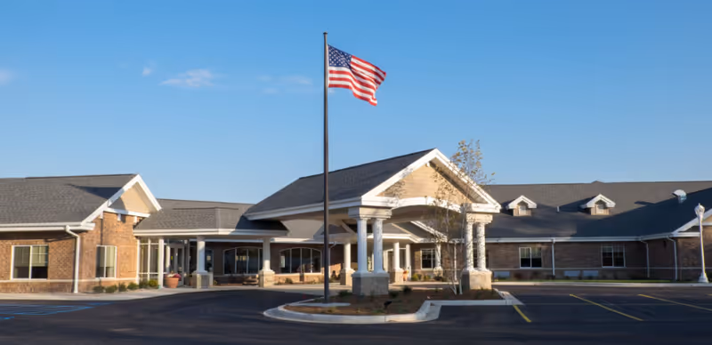 Front entrance of Breton Woods senior living facility with an American flag flying in front and a paved driveway.