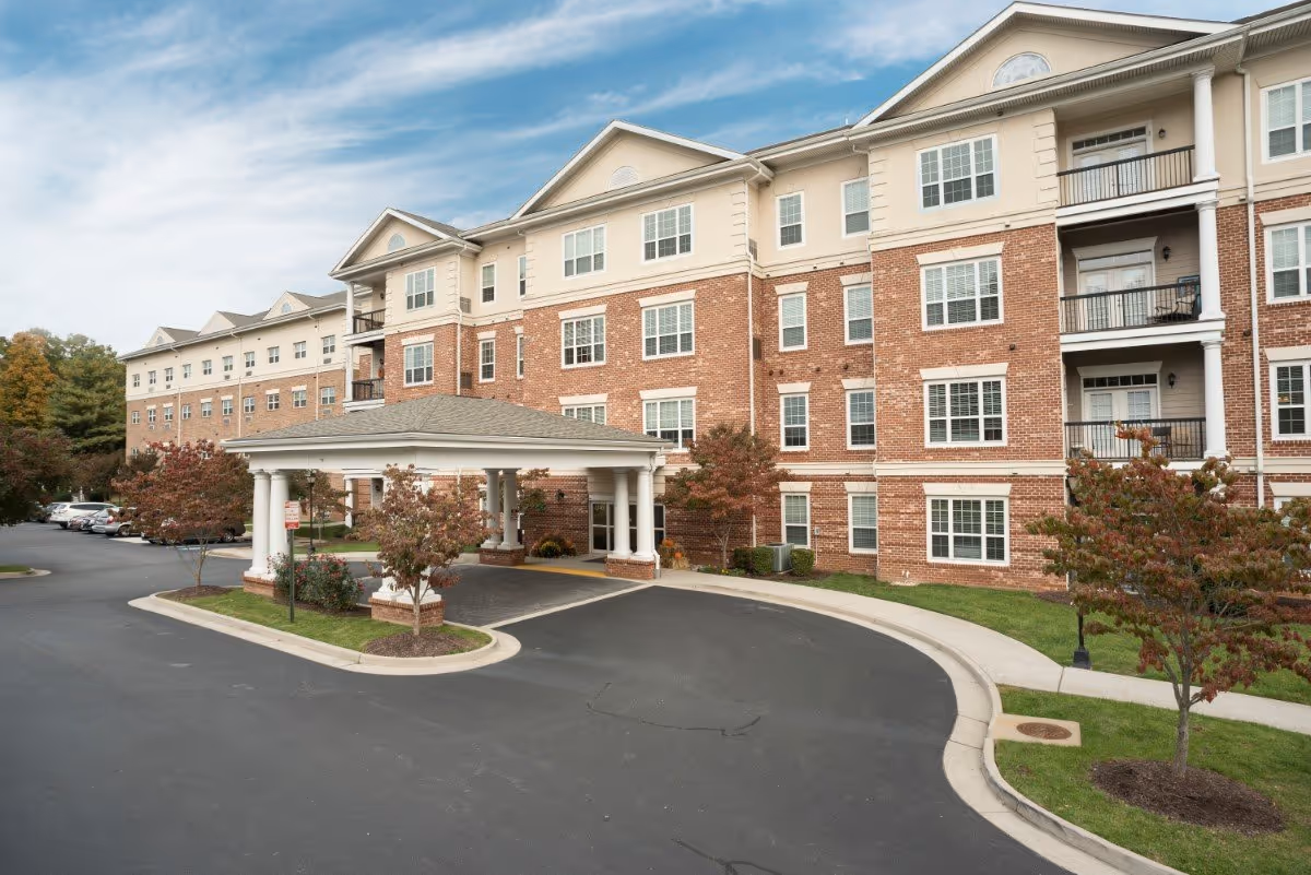 Front exterior of a multi-story brick senior living building with a covered entrance driveway and landscaped grounds.