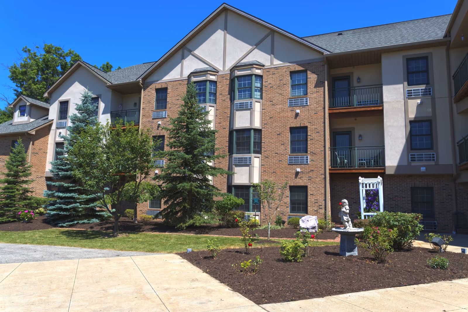 Three-story brick and stucco building with balconies, landscaped front garden, trees, and a small statue in the courtyard.