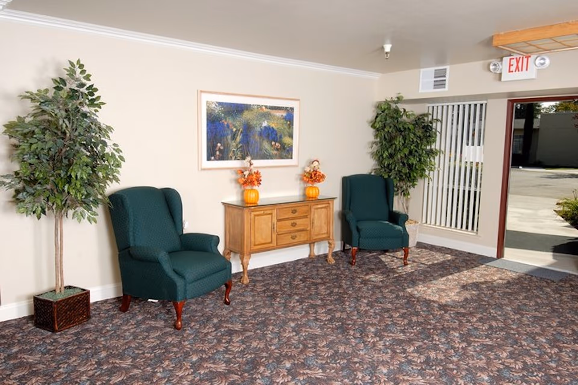 Lobby seating area with two green armchairs, a wooden console table with fall decorations, potted plants, and an open exit door.