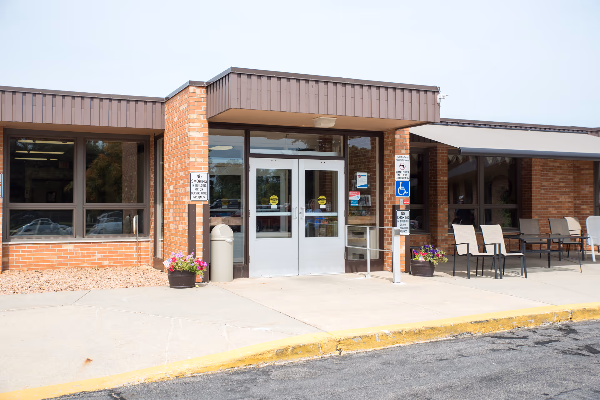 Exterior front entrance of a brick building with double glass doors, a small ramp with handrails, potted flowers, a trash can, and several chairs under an awning. Signs near the entrance indicate no smoking and handicap accessibility.
