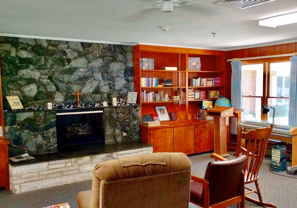 A cozy living room with a stone fireplace on the left, decorated with a small cross and candles. To the right of the fireplace is a wooden built-in bookshelf filled with books and framed pictures. In front of the fireplace are two chairs, one upholstered and one wooden. A large window with blue curtains lets in natural light, and a keyboard and globe are visible near the window.
