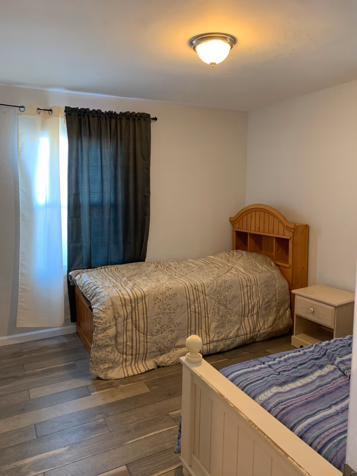 Two single beds with wood headboards, nightstand, and curtains in a bright bedroom.