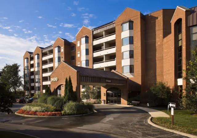 Exterior view of a multi-story brick building with balconies and a covered entrance. The building number 2600 is displayed on the entrance structure. There are landscaped bushes and flowers in front, with a driveway leading to the entrance under a partly cloudy sky.