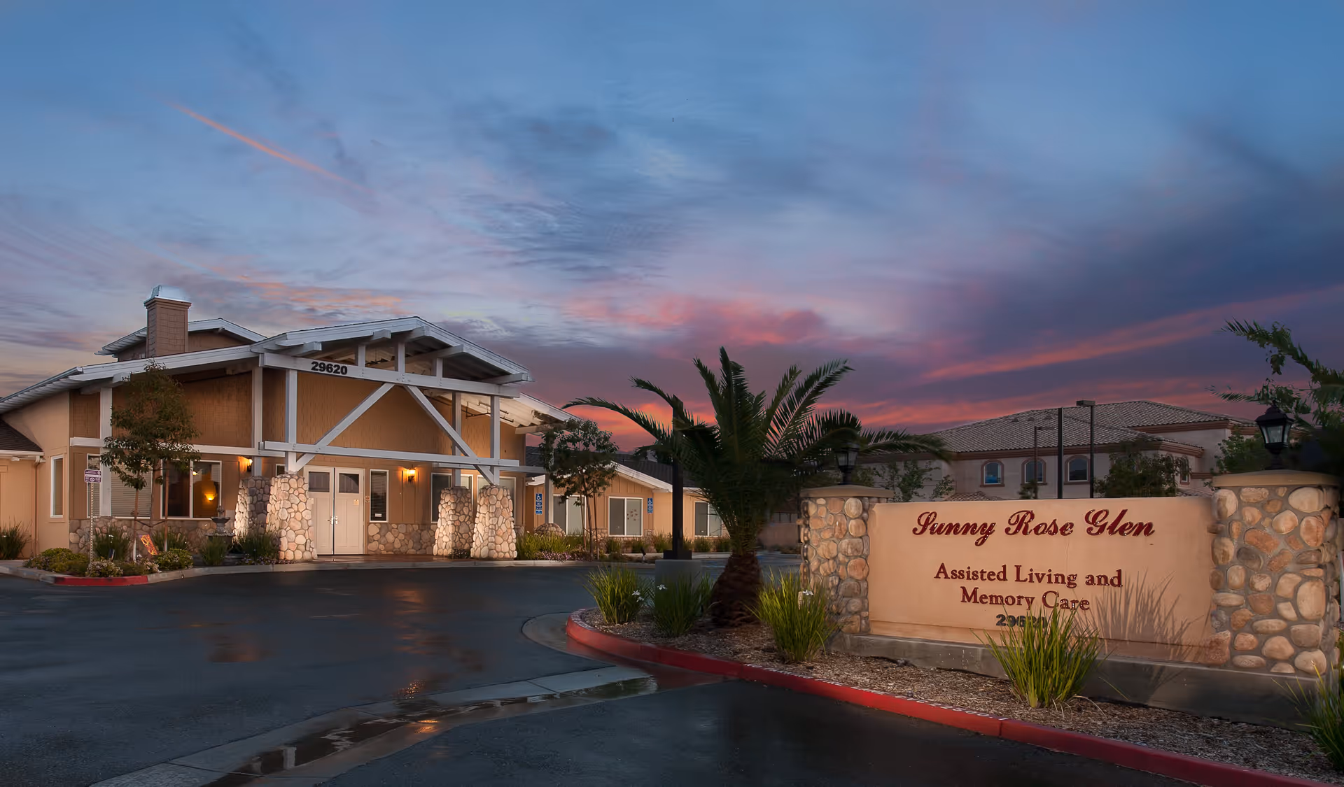 Exterior view of Sunny Rose Glen assisted living and memory care facility at dusk, showing the building entrance with stone pillars and a sign with the facility name and services.