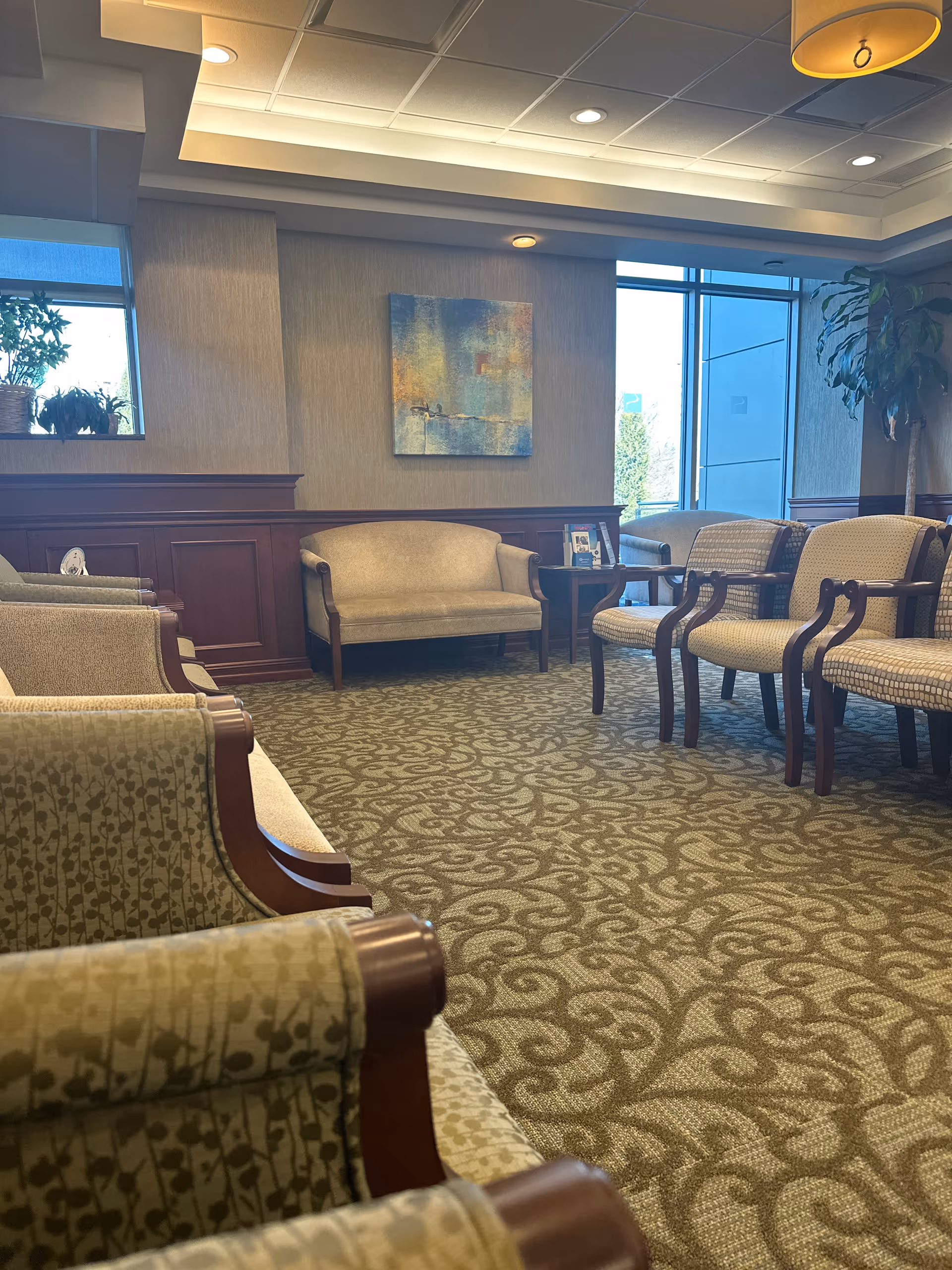 Spacious waiting lounge with upholstered seating, wood paneling, artwork, and large windows.