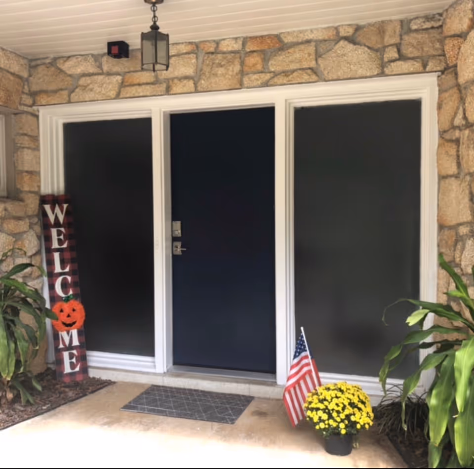Stone-front entrance with dark doors, a welcome sign, pumpkins, flag, and yellow mums.