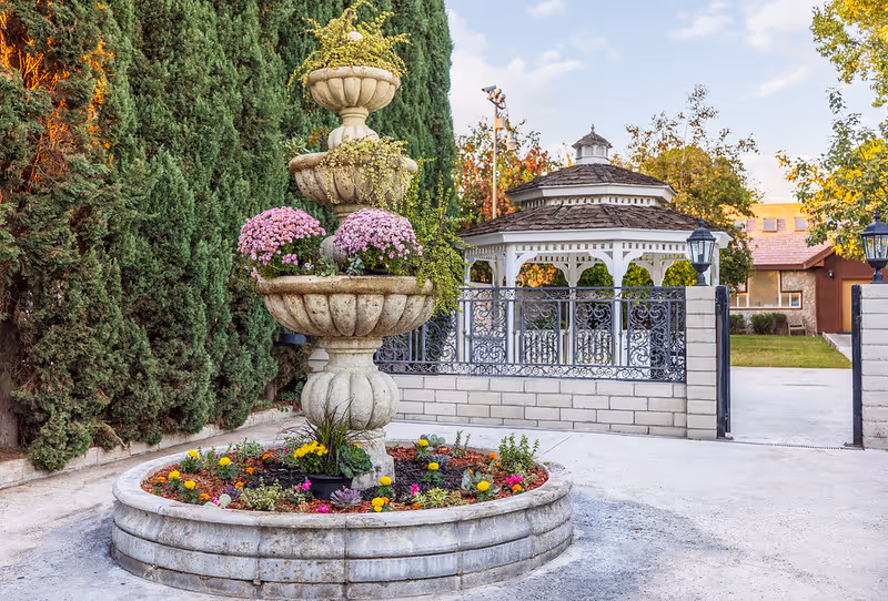 A tiered stone fountain decorated with pink flowers and greenery in the center of a circular flower bed with various colorful flowers. Behind the fountain, there is a black wrought iron gate and a white gazebo with a shingled roof. Tall green trees and a building with a brown roof are visible in the background under a partly cloudy sky.