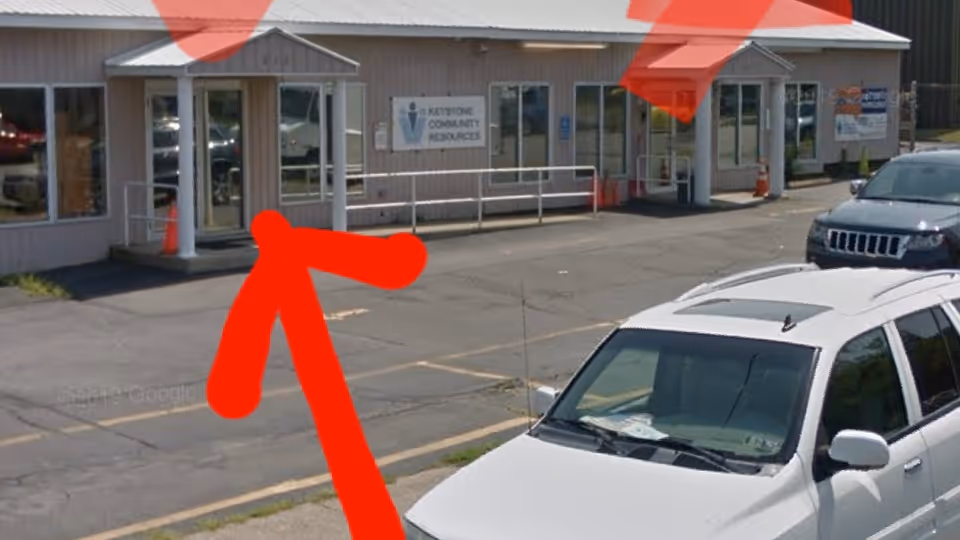 Exterior view of a small facility with glass entrances, ramp, and parked cars.