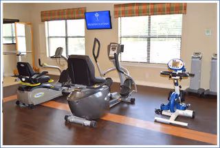 Exercise room with several cardio machines (recumbent bike, upright bike, and elliptical) in front of windows and a wall-mounted TV.