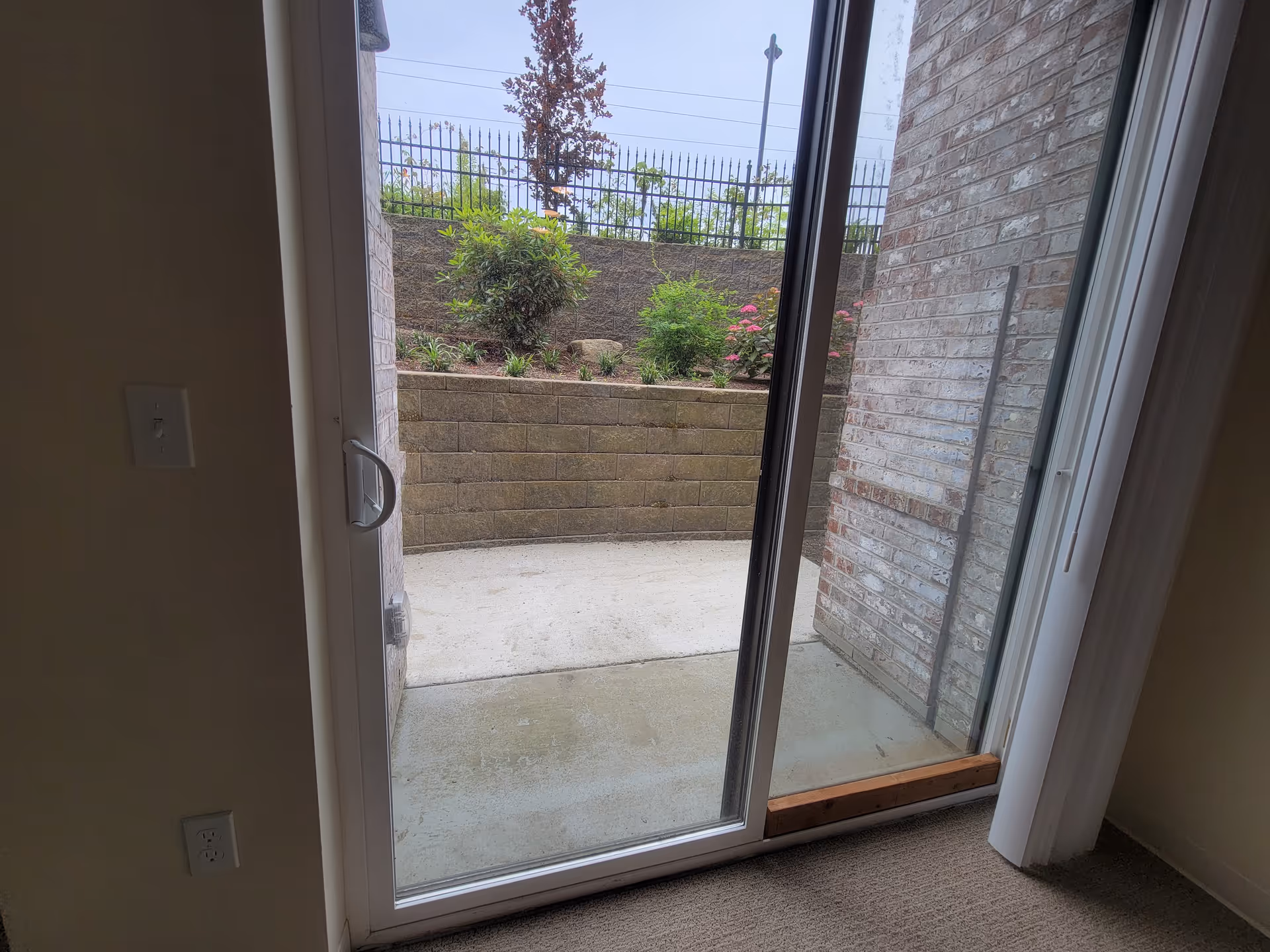 Open sliding glass door to a concrete patio with brick walls and shrubs.