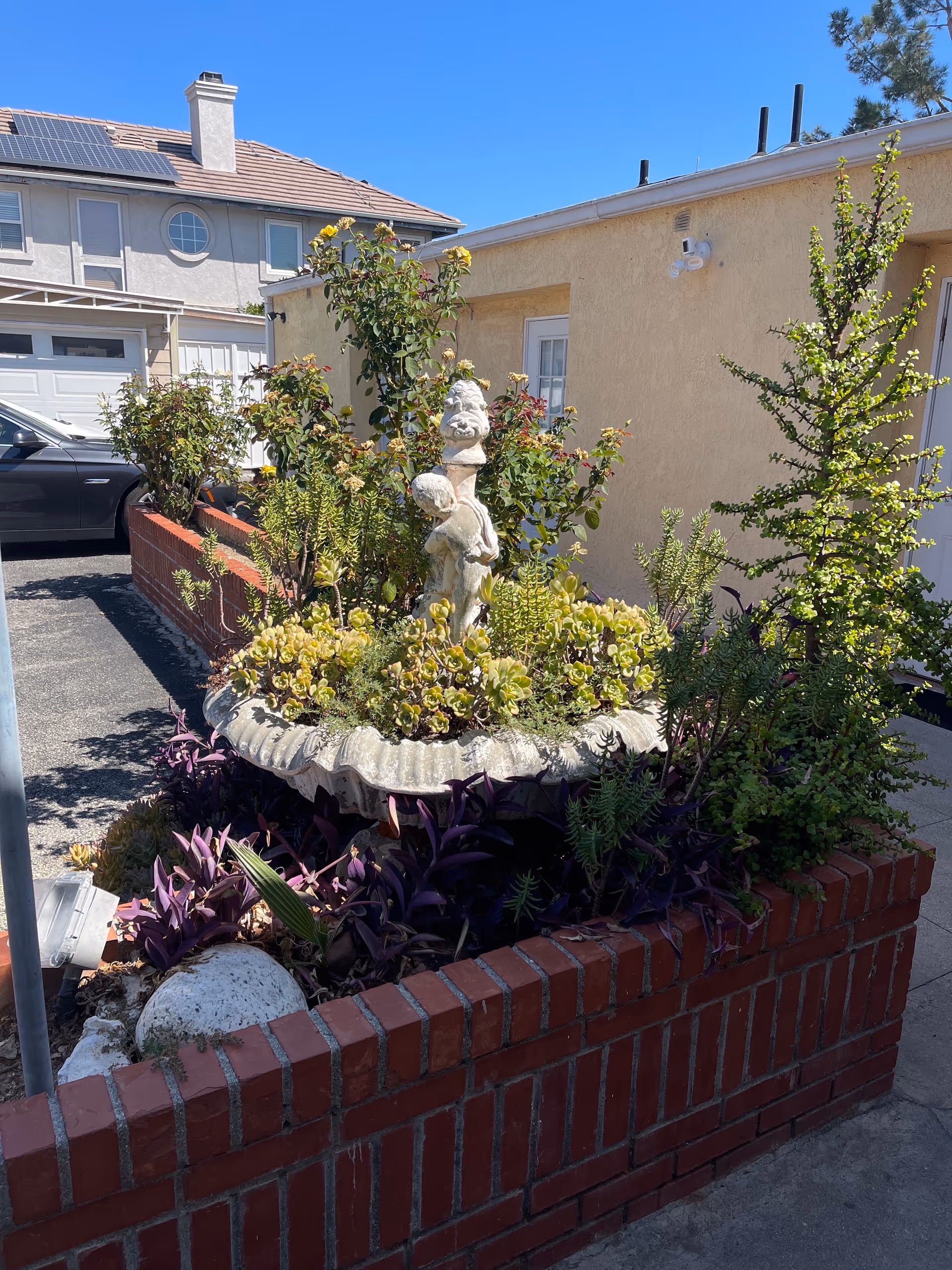 Outdoor brick planter with a statue and various plants beside a beige building.