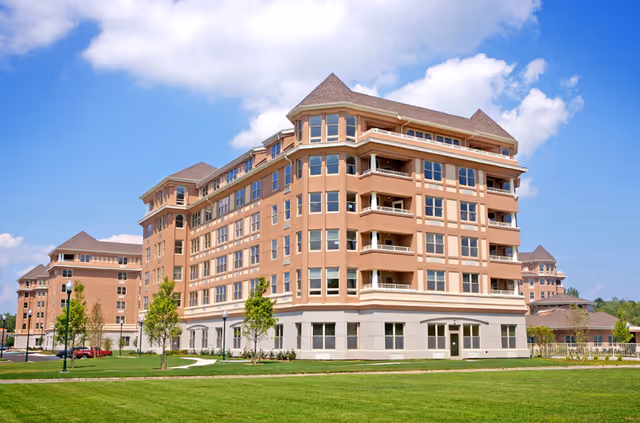 Large multi-story beige senior living building with balconies set on a grassy lawn under a blue sky.