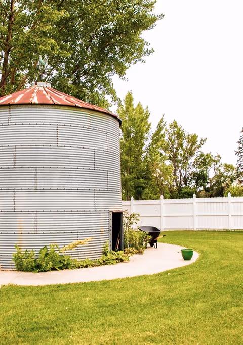 Corrugated metal silo in a grassy yard with a white fence, trees, a wheelbarrow and a green planter.