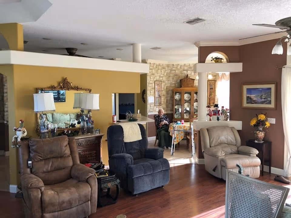 A cozy living room area with three recliner chairs in brown, dark blue, and beige colors. The room has wooden flooring and walls painted in warm tones of yellow and brown. There is a decorative mirror with two lamps on a wooden console table against the yellow wall. In the background, an elderly person is seated at a dining table covered with a floral tablecloth, near a cabinet displaying plates. The room is well-lit with natural light coming through a window with sheer curtains.