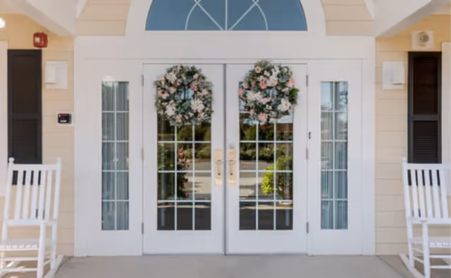Exterior entrance with double glass doors, floral wreaths, beige siding, and white rocking chairs.