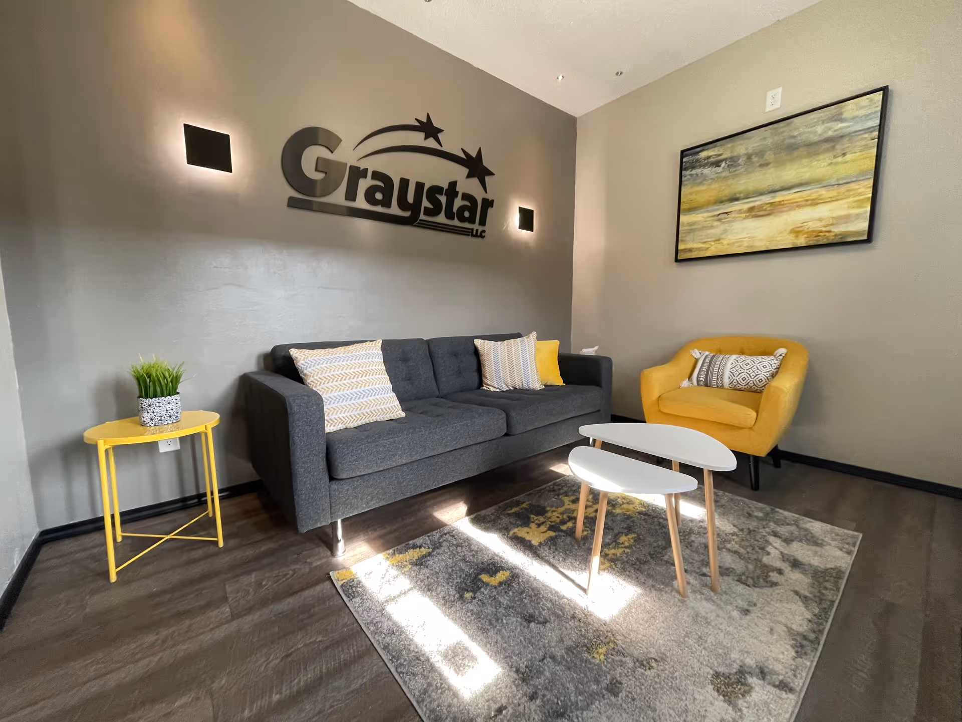 Modern lounge area with gray sofa, yellow chair, coffee tables, and Graystar logo wall art.