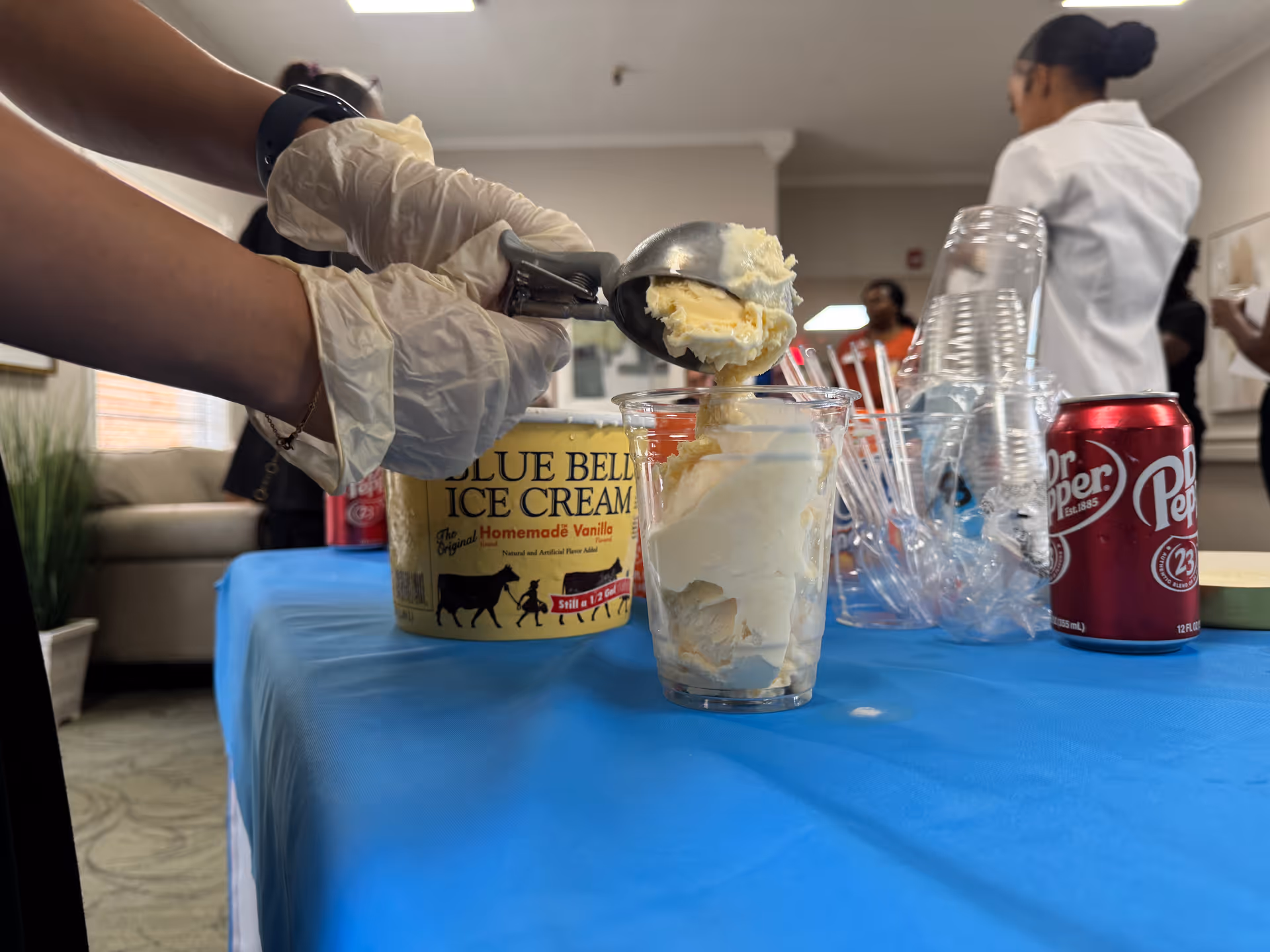 Hands scoop vanilla ice cream into a glass on a blue table at an indoor event.