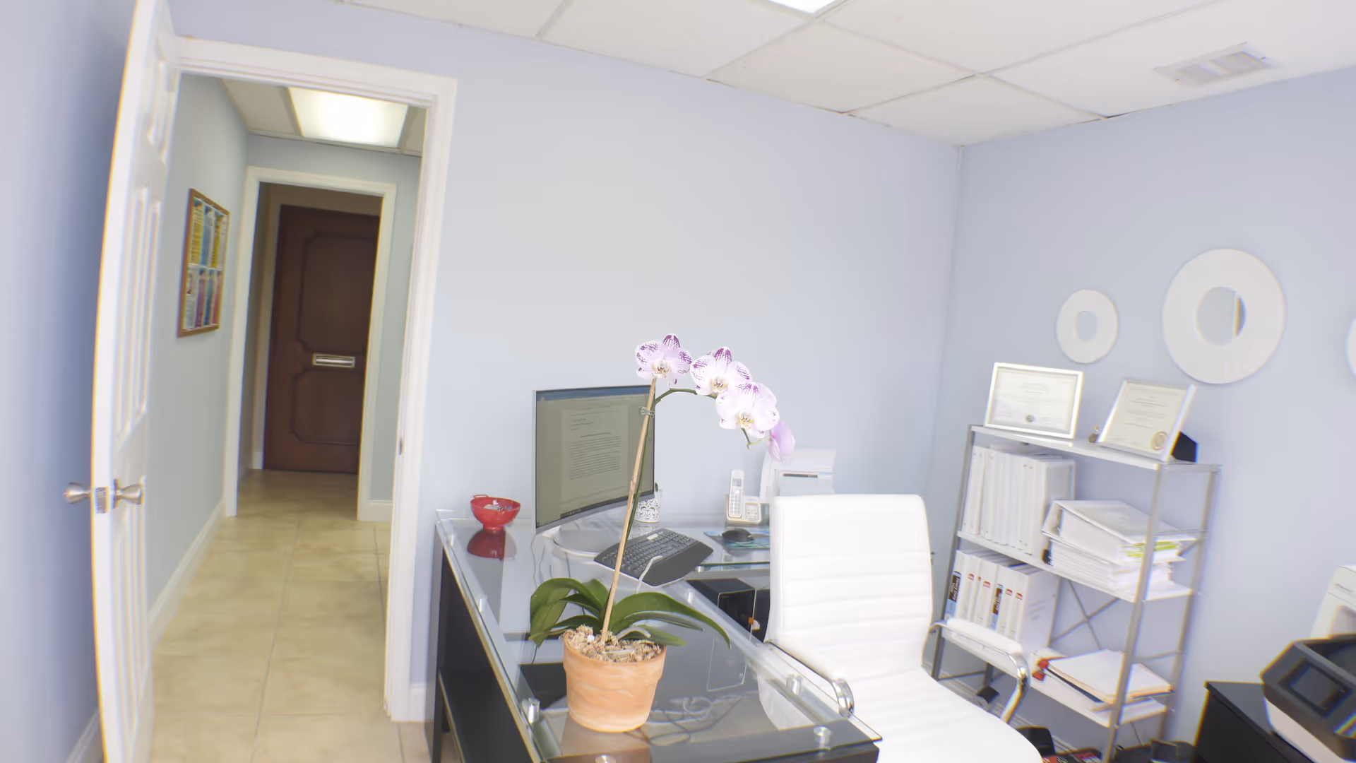 Counseling office space with glass desk, white chair, orchid plant, and shelves.