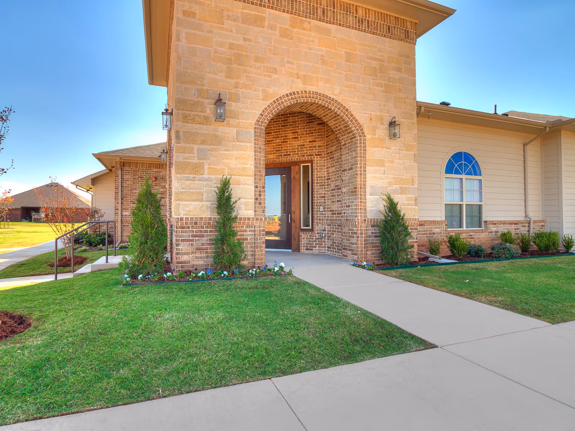 Front entrance of a building with a stone and brick archway, a glass door, and outdoor wall lanterns. The building is surrounded by green grass, small shrubs, and a concrete walkway leading to the door under a clear blue sky.