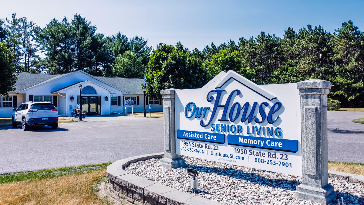 Exterior view of Our House Senior Living facility in Wisconsin Dells, showing a white building with a gray roof surrounded by trees. In the foreground, there is a large sign with the facility's name, services offered (Assisted Care and Memory Care), address, phone numbers, and website. A parked car is visible near the building entrance.