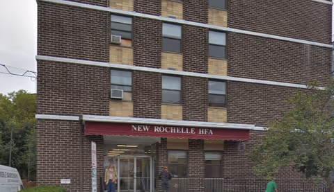 Exterior view of a multi-story brick building with a maroon awning above the entrance that reads 'NEW ROCHELLE HFA'. There are several windows with air conditioning units, a tree on the right side, and a few people near the entrance.