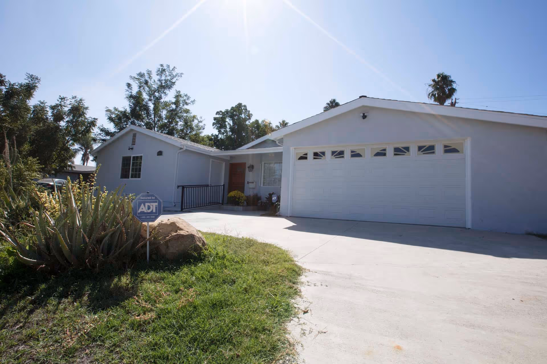Exterior view of a single-story residential building with a large white garage door, a front entrance with a wooden door, and a small garden area with plants and a rock. The sky is clear and sunny.