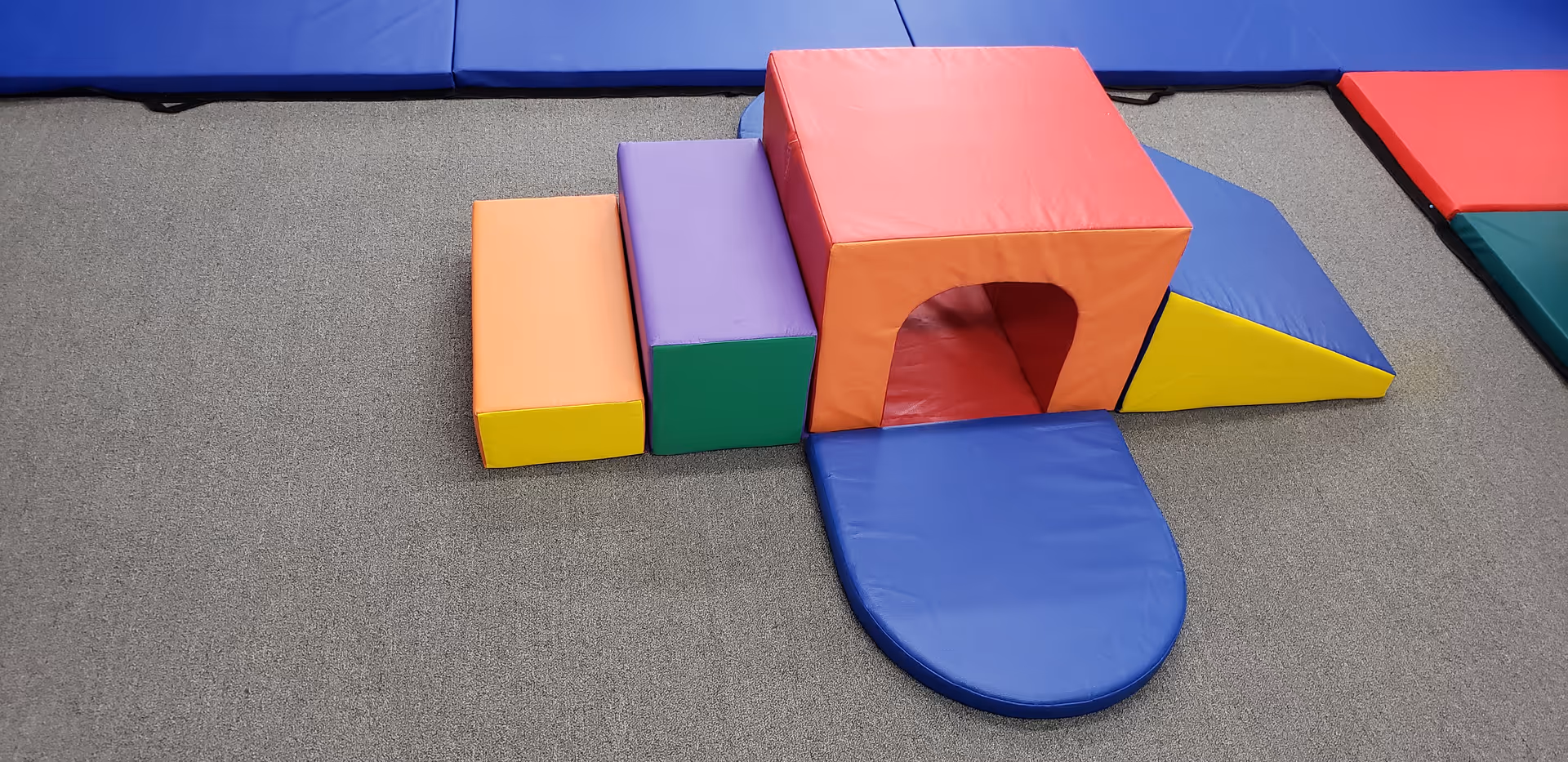 Colorful foam play blocks and a tunnel sit on a gray carpet in a rehabilitation space.