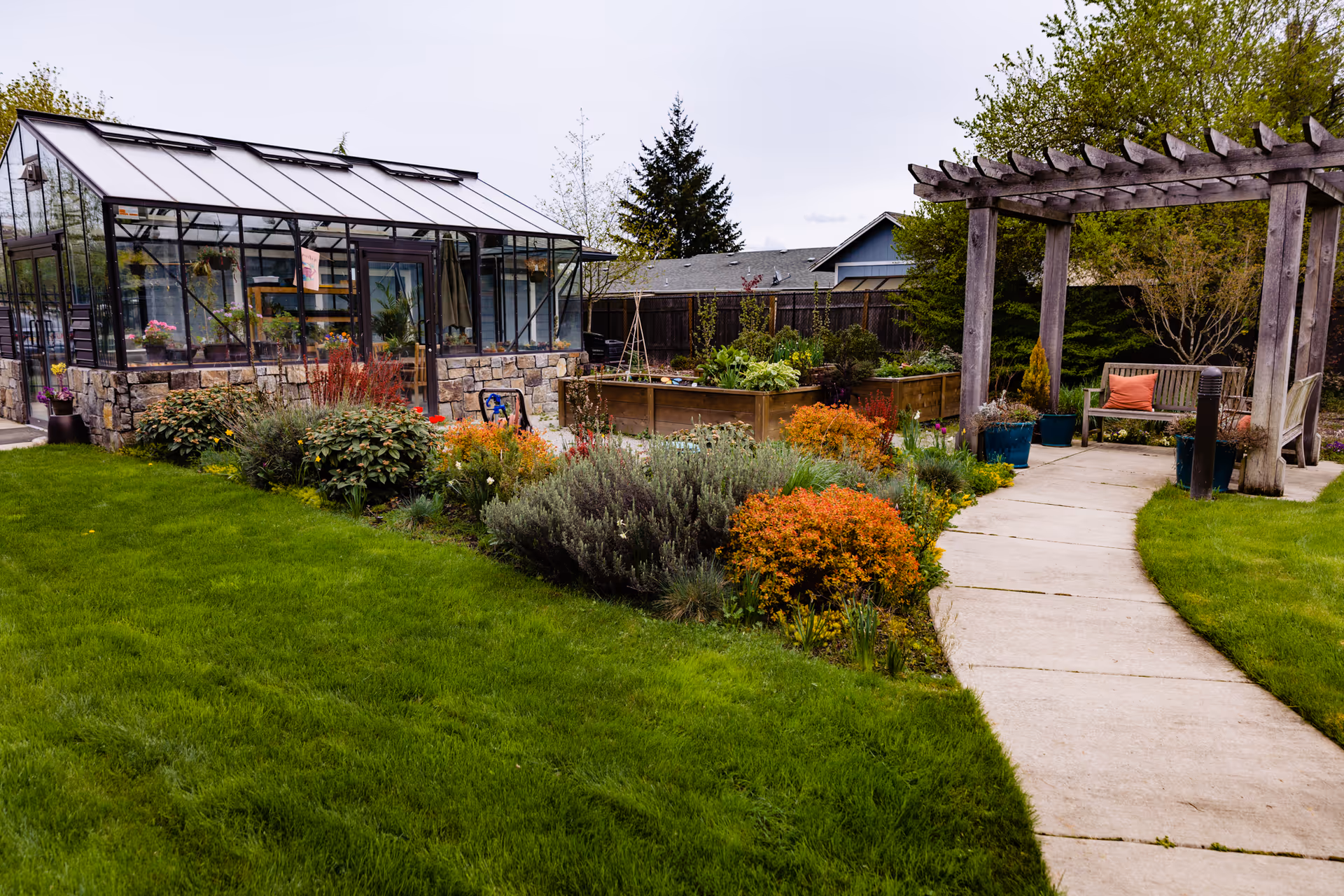 A landscaped senior living outdoor garden with a glass greenhouse, pergola, benches, and path.