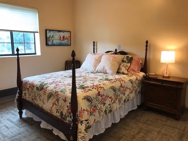 A well-lit bedroom with a four-poster bed covered in floral bedding, a wooden nightstand with a lamp, and a window.