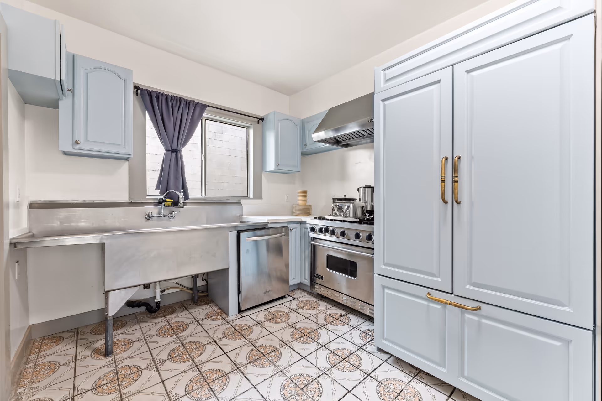 Blue kitchen boasting stainless steel sink, range, dishwasher, large cabinets, and a window.