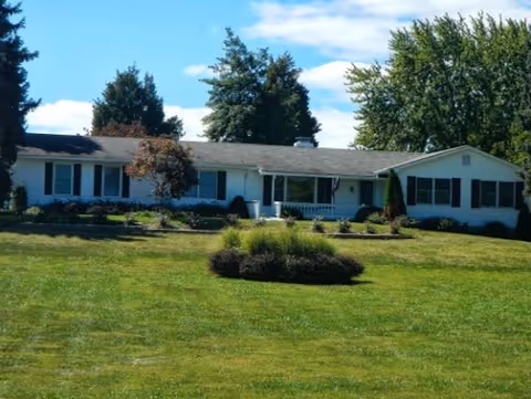 Single-story white residential building with a gray roof surrounded by green grass and trees under a partly cloudy blue sky.