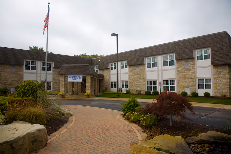 Exterior view of a two-story senior living facility building with stone and beige siding, multiple windows, a covered entrance with a sign reading 'Deficiency Free Facility,' a flagpole with an American flag, landscaped garden beds, and a brick-paved walkway leading to the entrance.