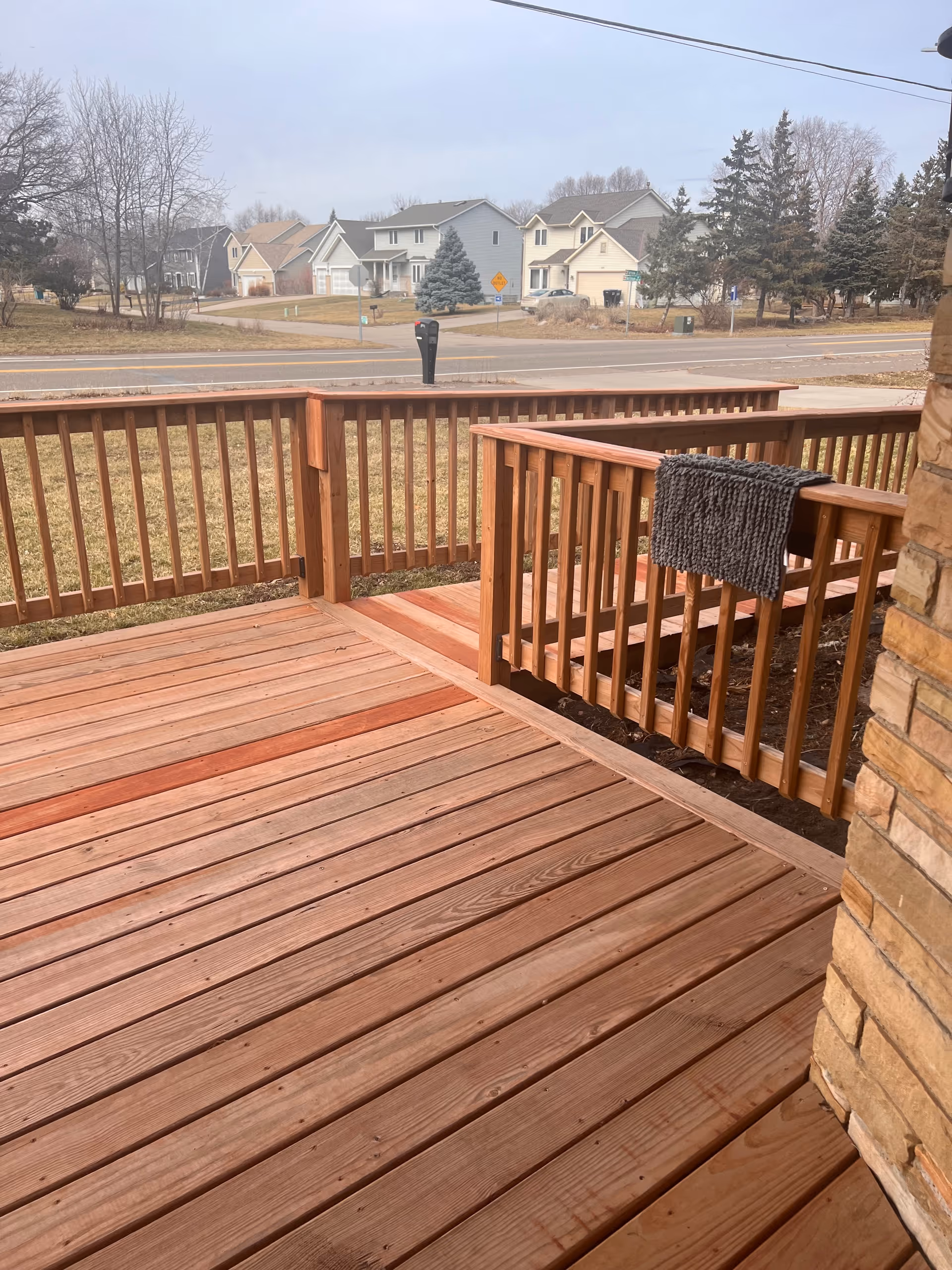 Wooden backyard deck with railing extends from a brick house, neighborhood street in background.