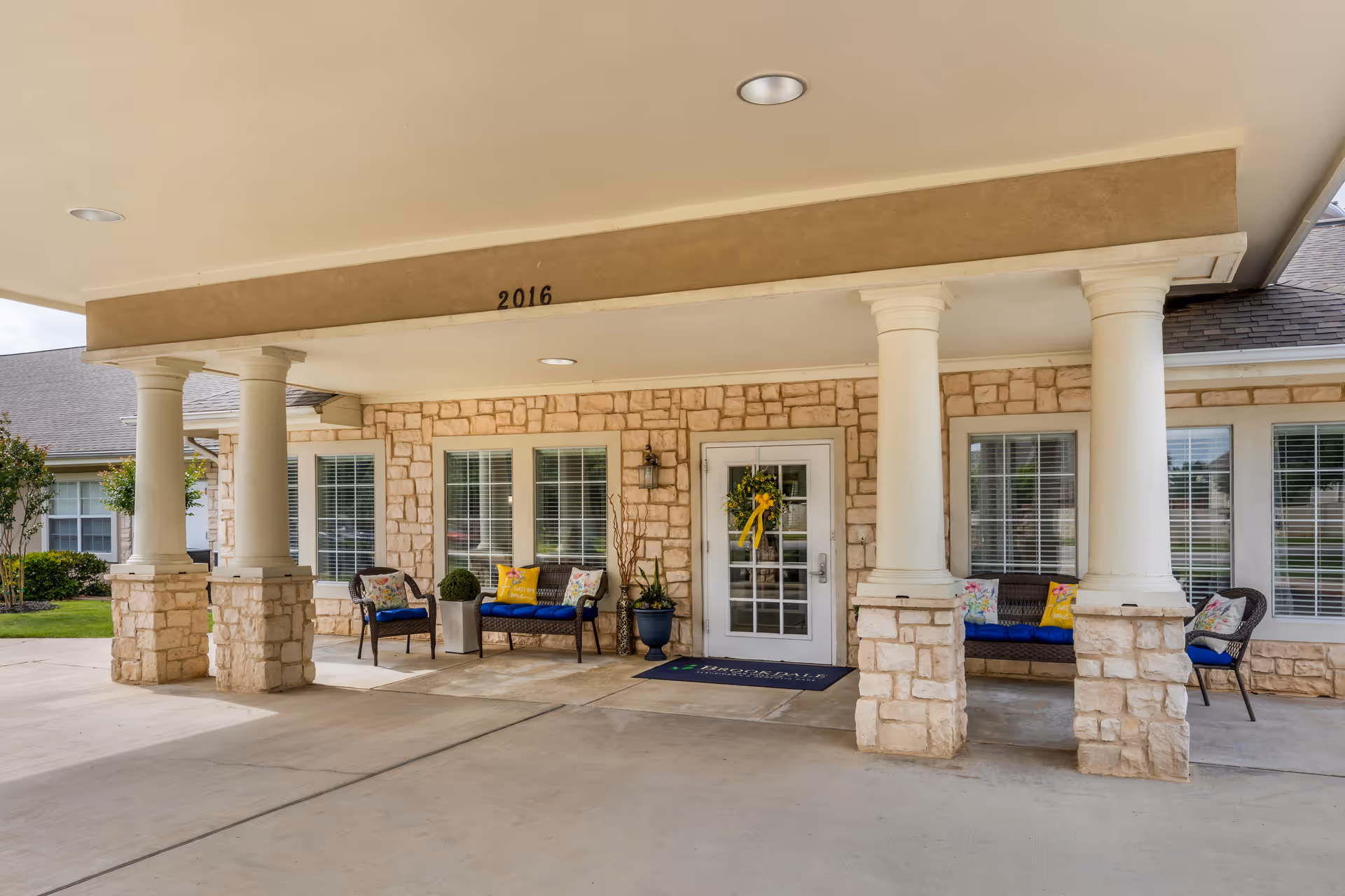 Covered entrance with stone columns, seating area, and glass door at Brookdale Oak Hollow.