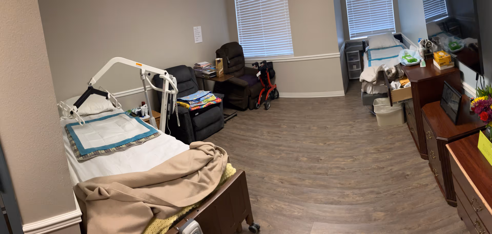 Two resident beds in a shared patient room with medical equipment, chairs, and a dresser.