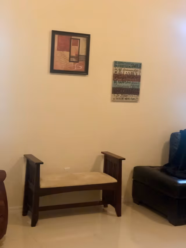 A small cushioned wooden bench against a beige wall with two framed artworks above and part of a leather chair at right.