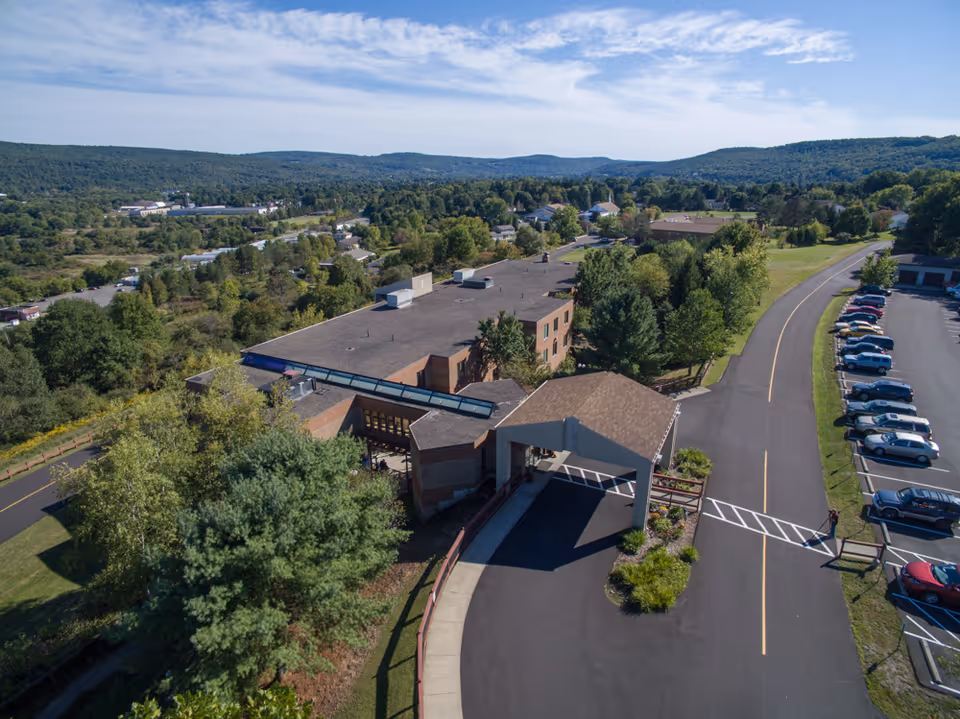 Aerial view of Norwich Rehabilitation & Nursing Center showing the main building with a covered entrance, surrounded by trees and greenery. A parking lot with several cars is visible to the right, and a winding road leads up to the facility. Hills and a partly cloudy sky are in the background.