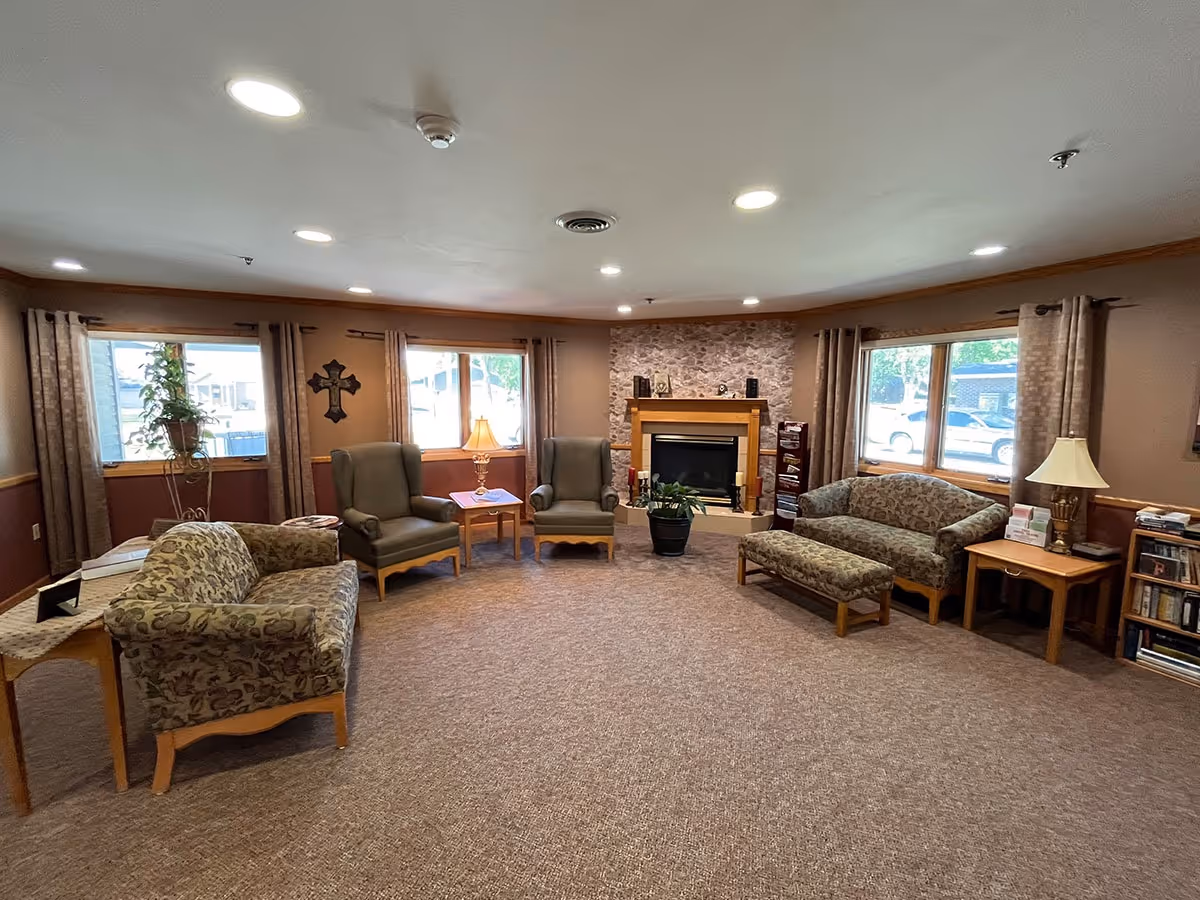 Cozy lounge area with couches, chairs, a fireplace, and large windows in a senior living facility.