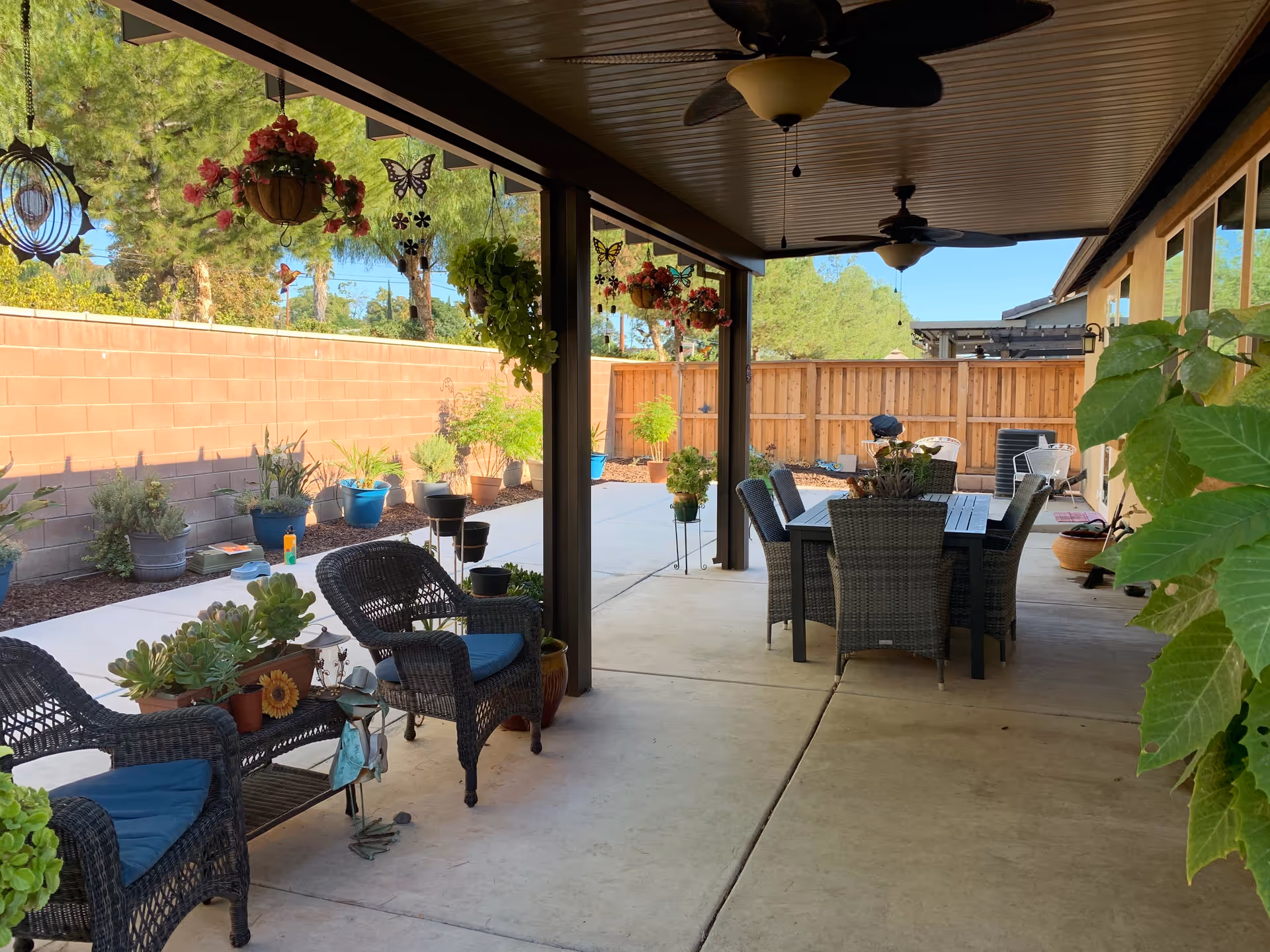 Covered outdoor patio with wicker seating, a dining table, hanging plants, and ceiling fans.