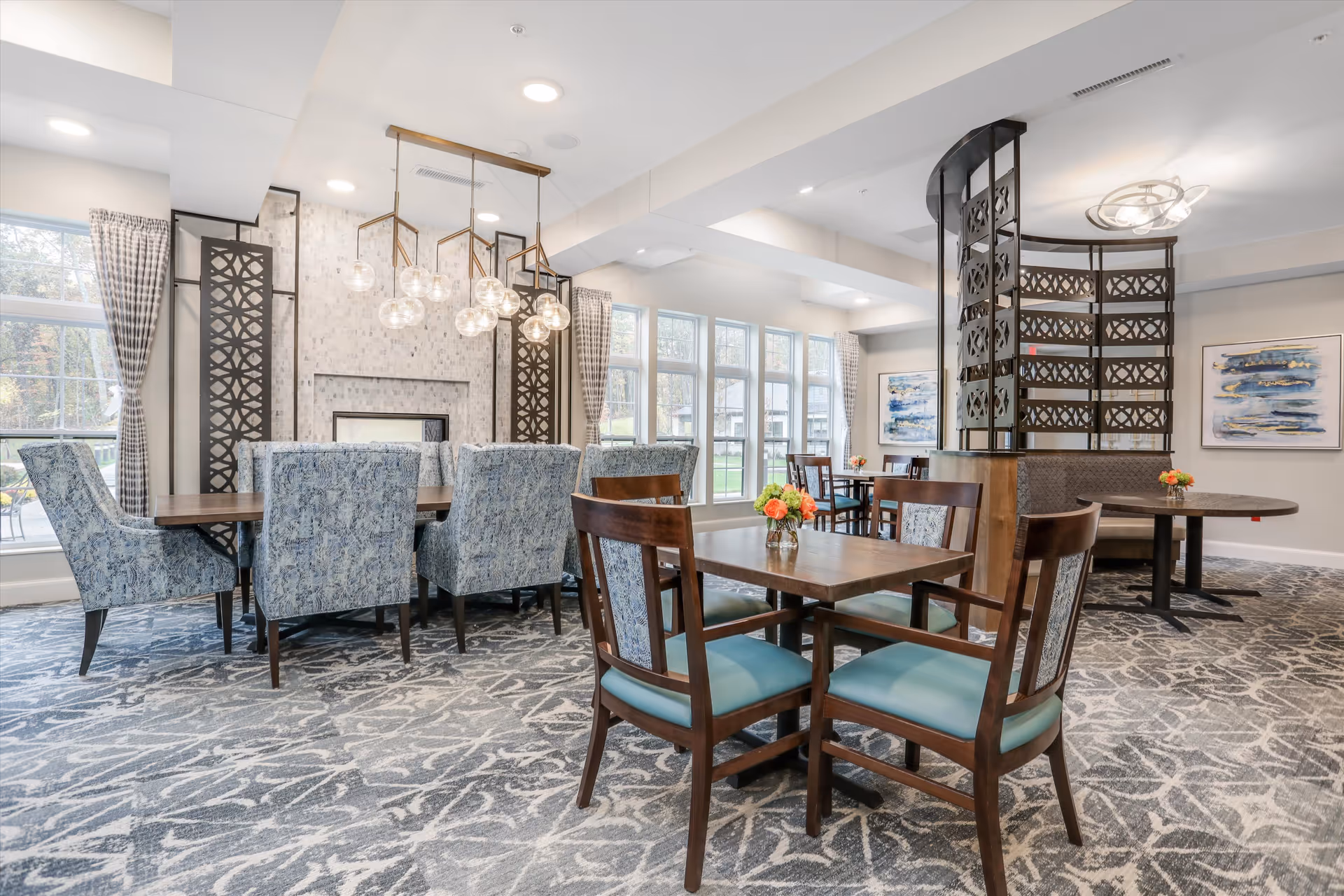 A bright and spacious dining area in Monark Grove Clarkston featuring multiple wooden tables with cushioned chairs. The room has large windows with checkered curtains allowing natural light to fill the space. There is a decorative fireplace with a modern light fixture hanging above a long table surrounded by blue patterned chairs. The carpet has a gray and white abstract pattern, and there are colorful floral centerpieces on the tables. The room also includes artistic wall decorations and a curved partition with intricate metal designs.