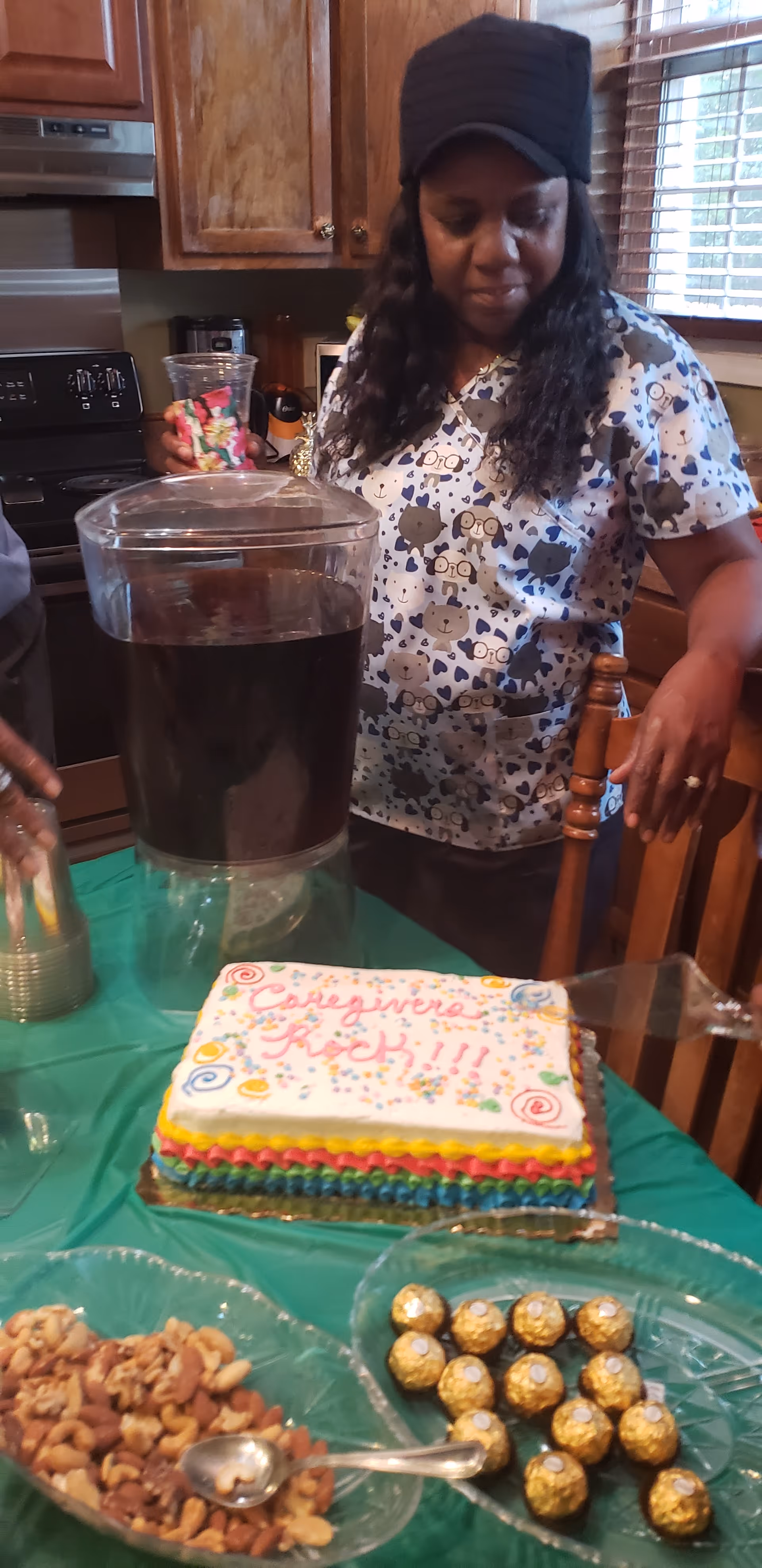 A woman wearing a patterned scrub top and a black cap stands next to a table with a colorful cake that says 'Caregivers Rock!!!', a large beverage dispenser filled with a dark drink, a bowl of mixed nuts, and a bowl of wrapped chocolates. The setting appears to be a kitchen with wooden cabinets and a window with blinds.