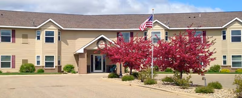 Two-story beige senior living facility with entrance, flagpole, and pink flowering trees.