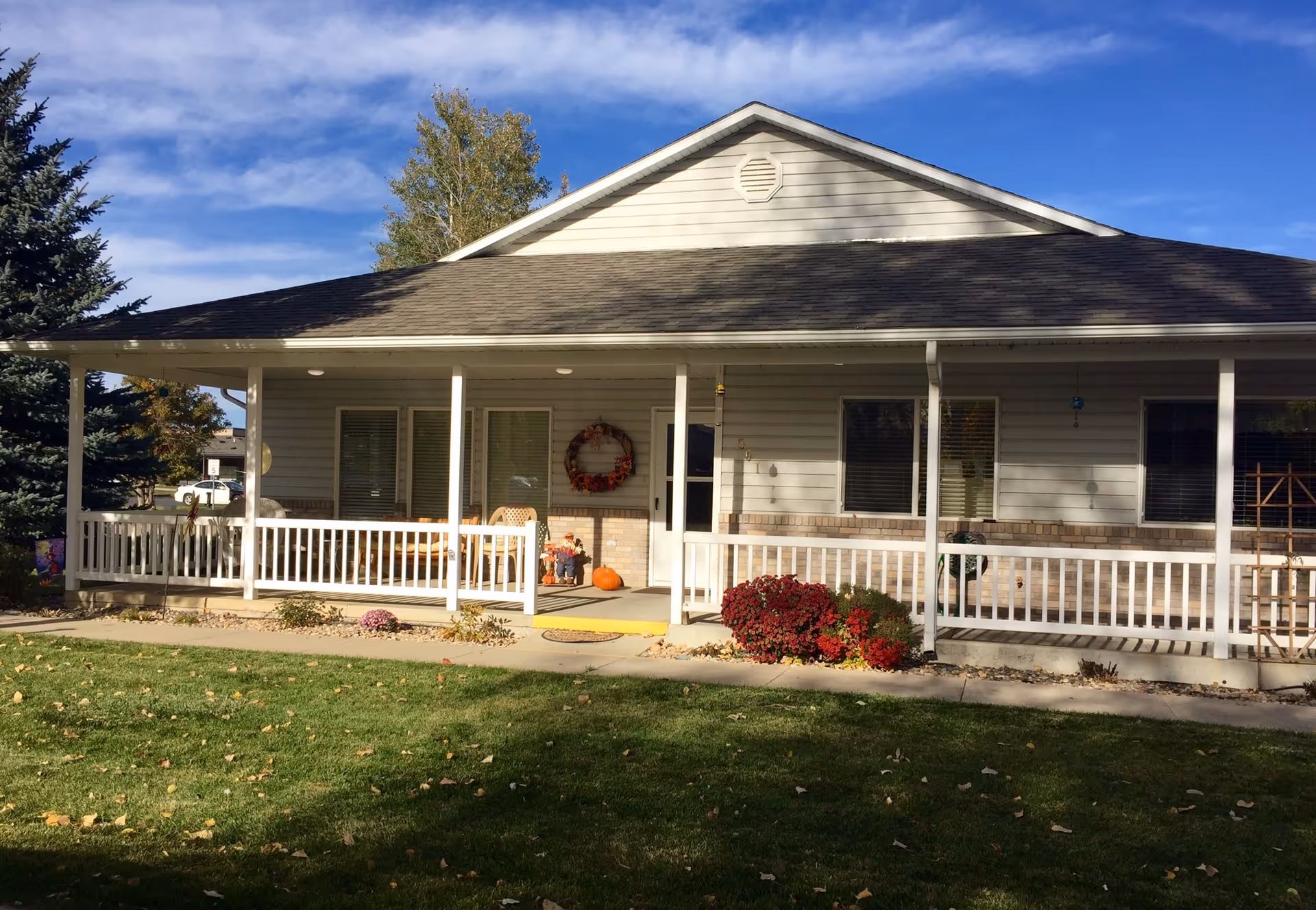 Front exterior view of a single-story building with a covered porch, white railing, and a few chairs. There are autumn decorations including a wreath on the door and pumpkins on the porch. The building is surrounded by green grass, some bushes with red flowers, and trees under a blue sky with some clouds.