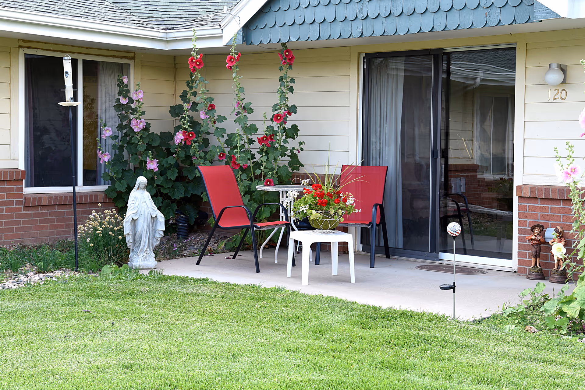 Outdoor patio with two red chairs and a small table holding a flower arrangement in front of a sliding glass door, surrounded by hollyhocks, a lawn, and a statue.