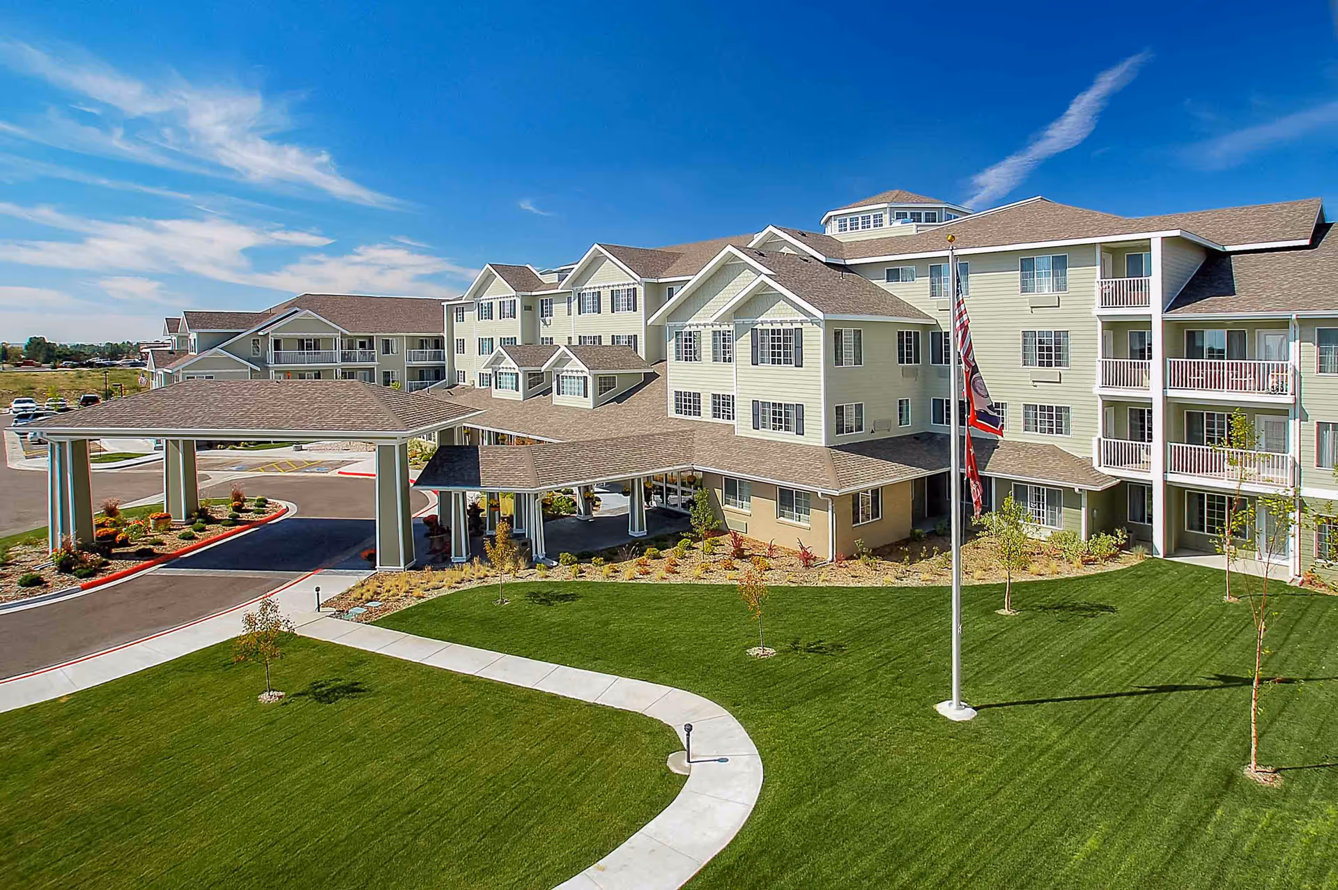 Large multi-story senior living building with a covered porte-cochere, flagpoles, and manicured lawn under a blue sky.