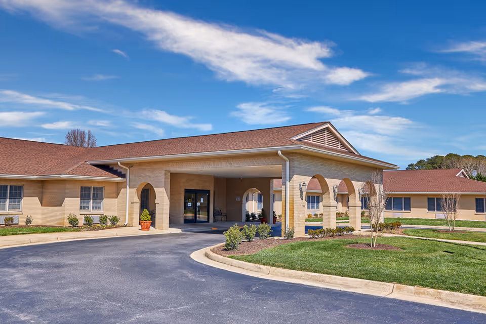 Single-story brick senior living facility with a covered entrance, arched columns and a circular driveway under a blue sky.