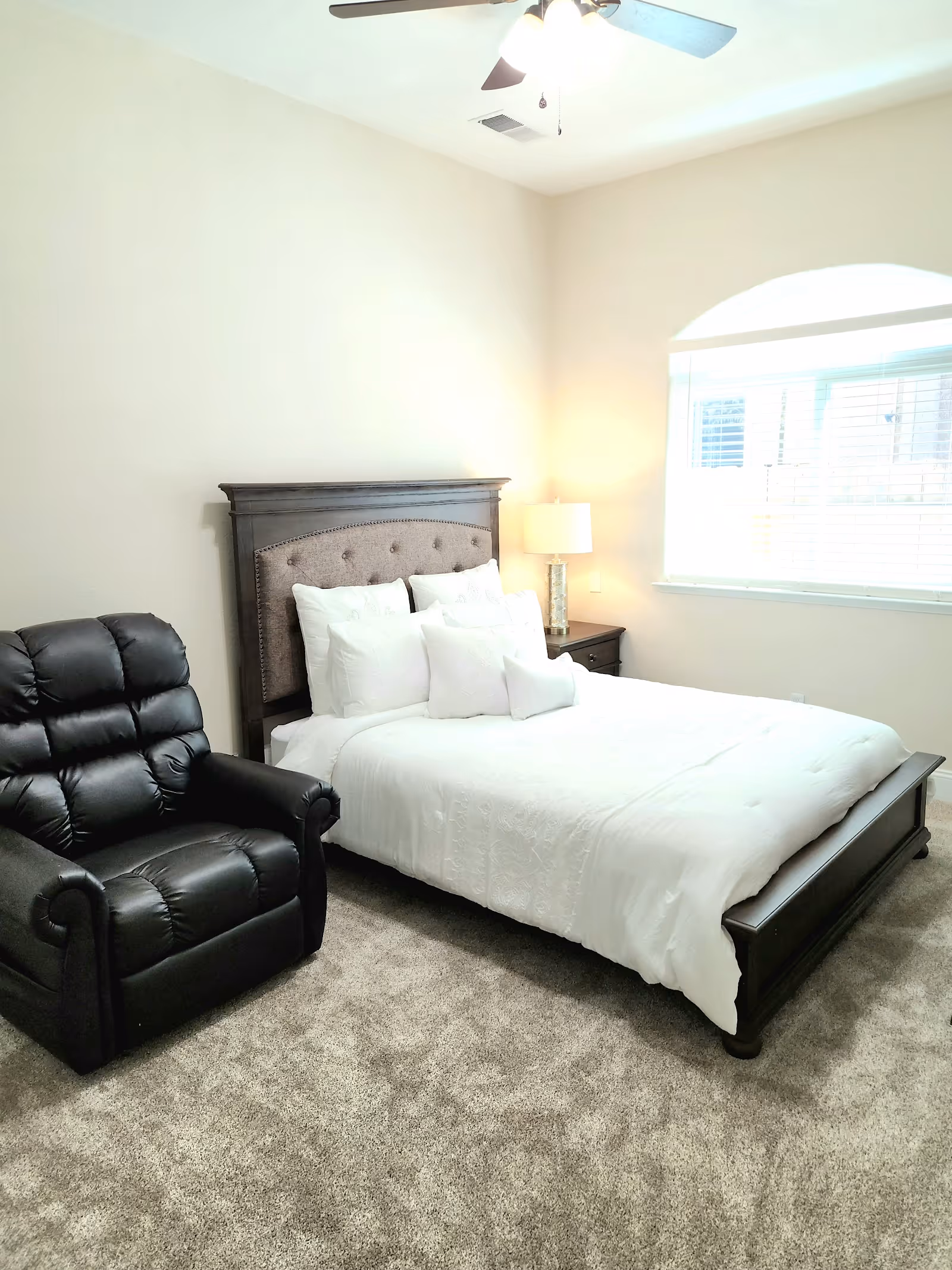 A bedroom with a white comforter, dark wood bed, nightstand, lamp, and black recliner.